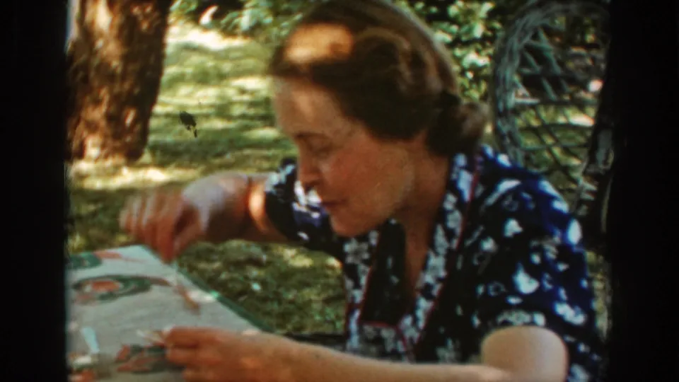 The woman leans down over her plate, using a utensil at the picnic table beneath trees and mottled sunlight.