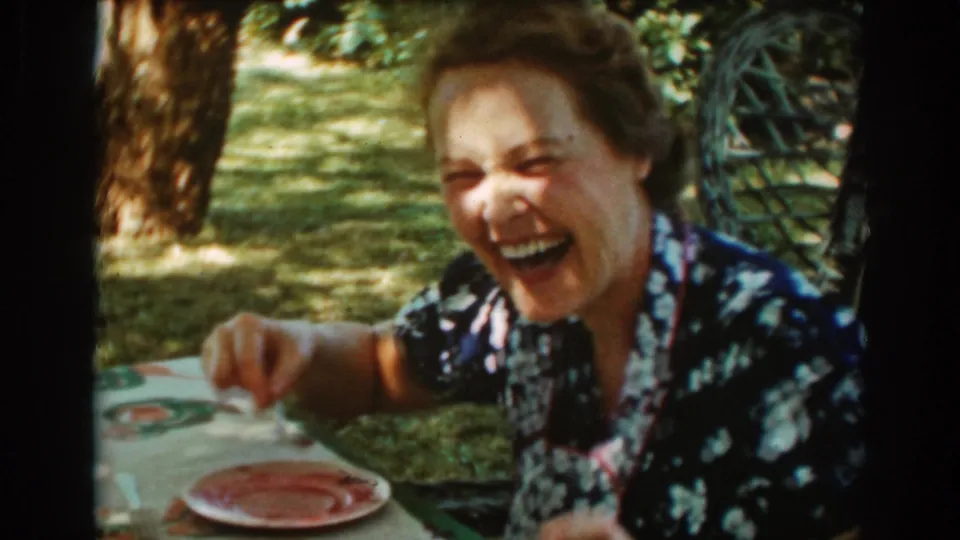 A close home-movie frame shows the woman laughing toward the camera while seated at the garden picnic table.