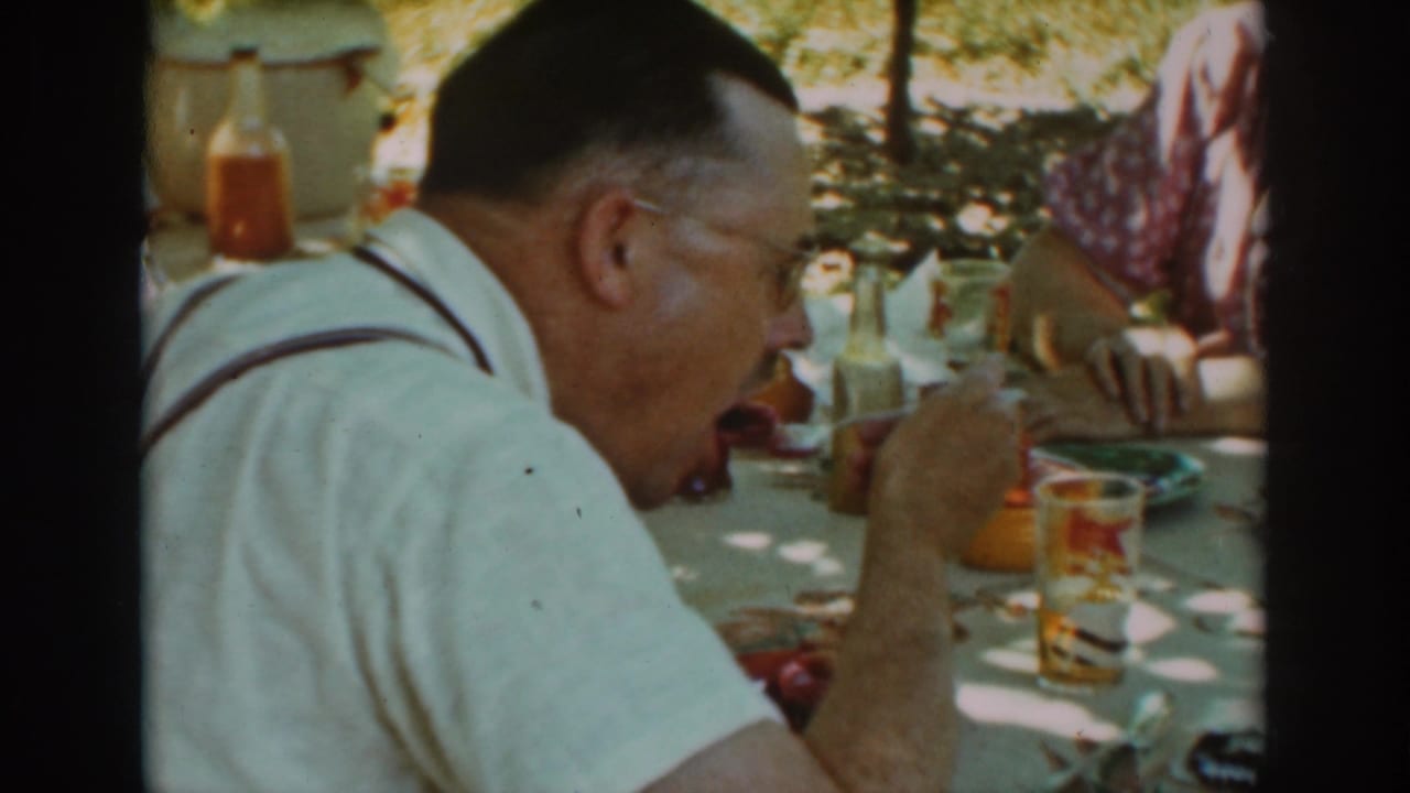 Man in suspenders eating at an outdoor table with glasses and bottles nearby.