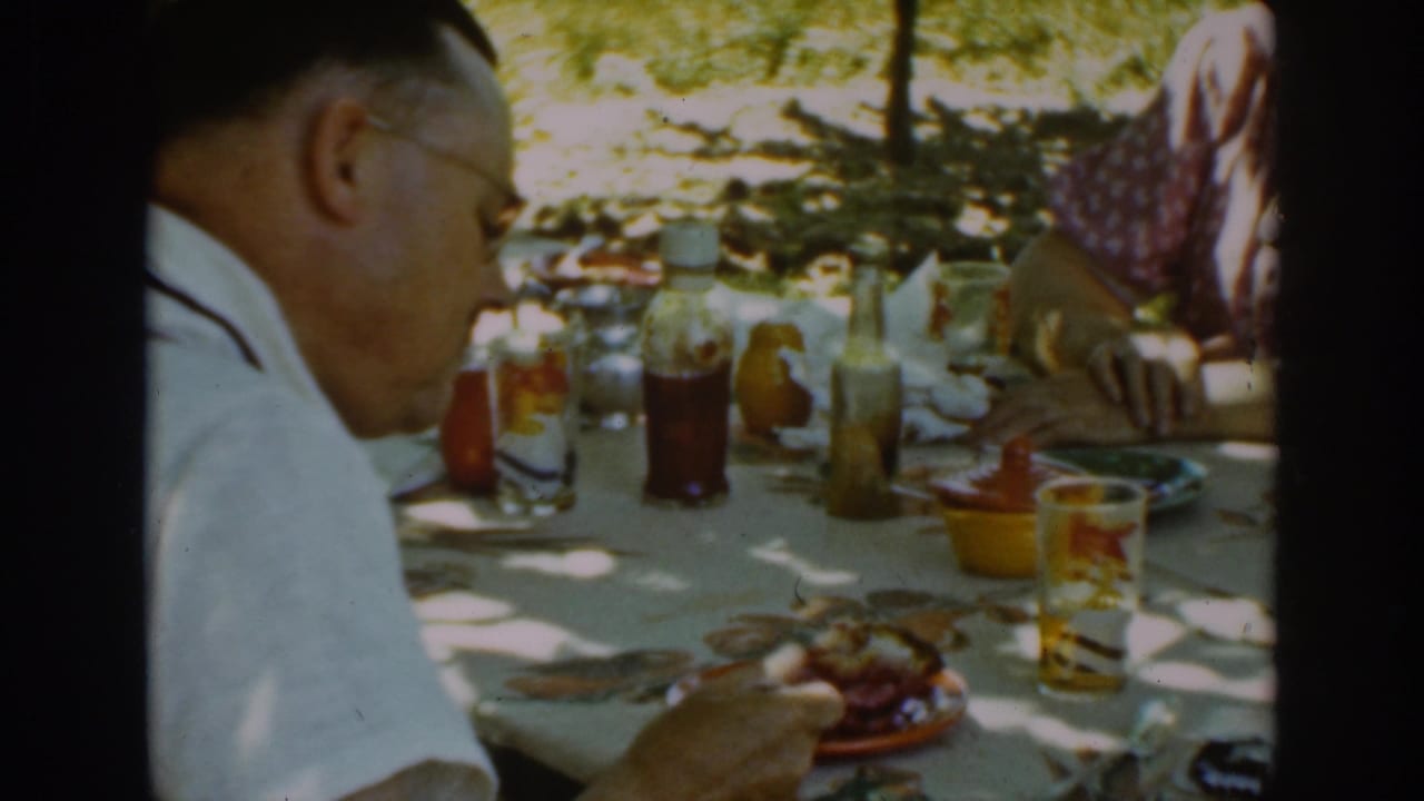 Man in a white shirt and suspenders sits at an outdoor picnic table with food and drinks.