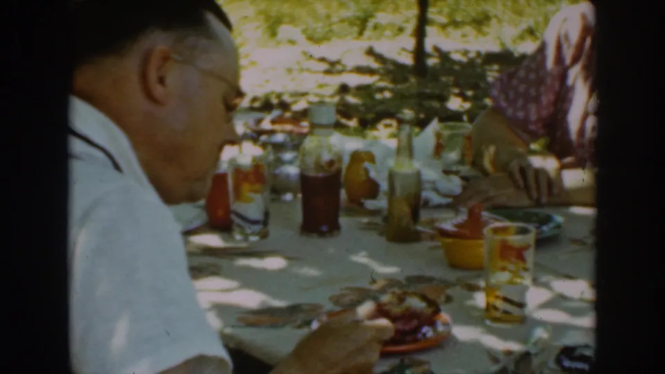 In 1939 Auburn, California home-movie footage, a man in glasses sits at a shaded picnic table with plates, glasses, and condiments.