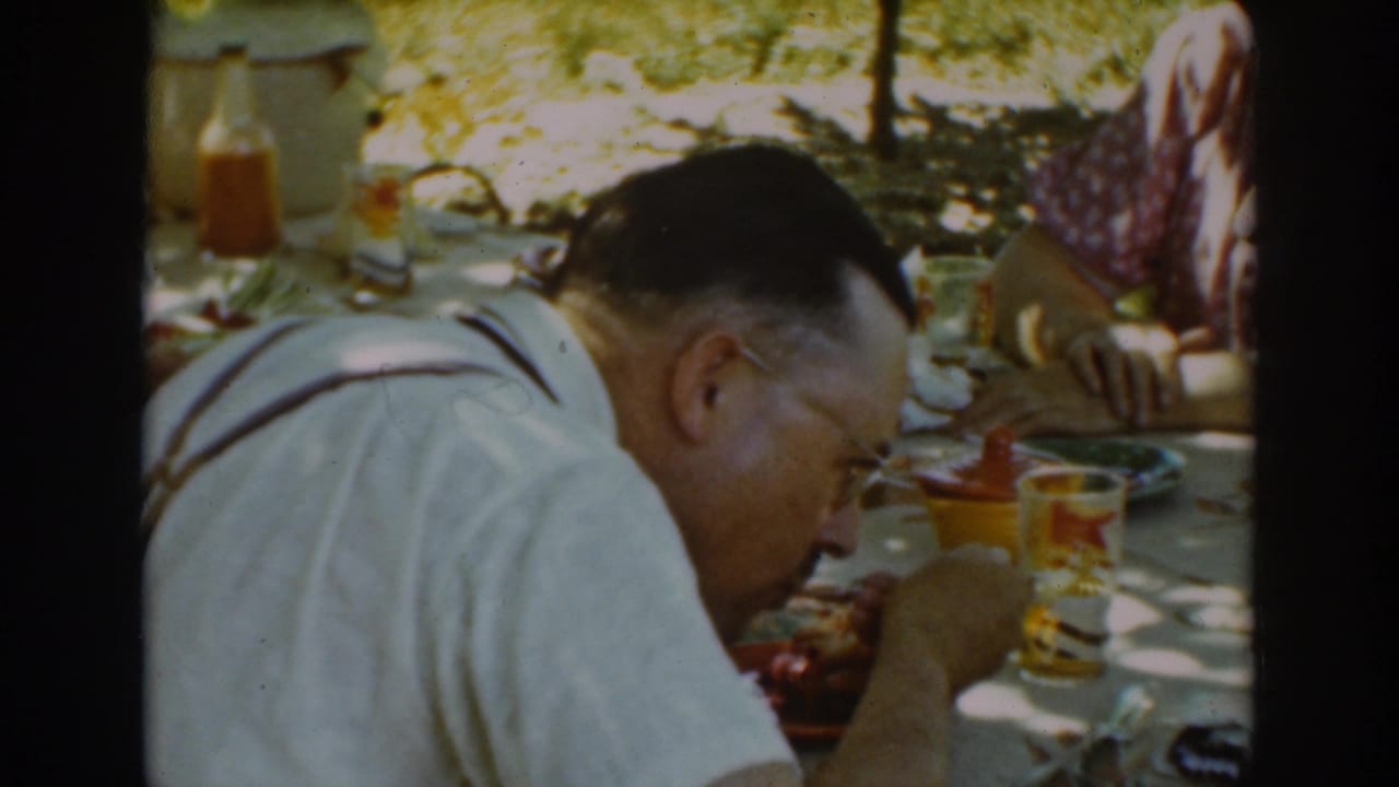 The man leans over his plate during a 1939 Auburn, California picnic, with drinks, condiments, and another adult partly visible across the table.