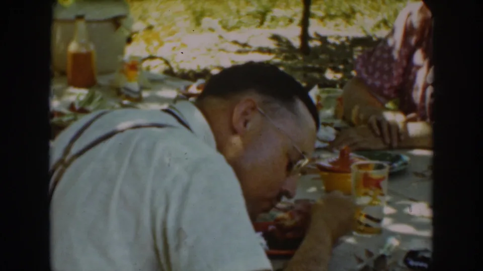 A close home-movie view shows the man eating at the shaded Auburn picnic table, with glassware, plates, and a partially visible companion nearby.
