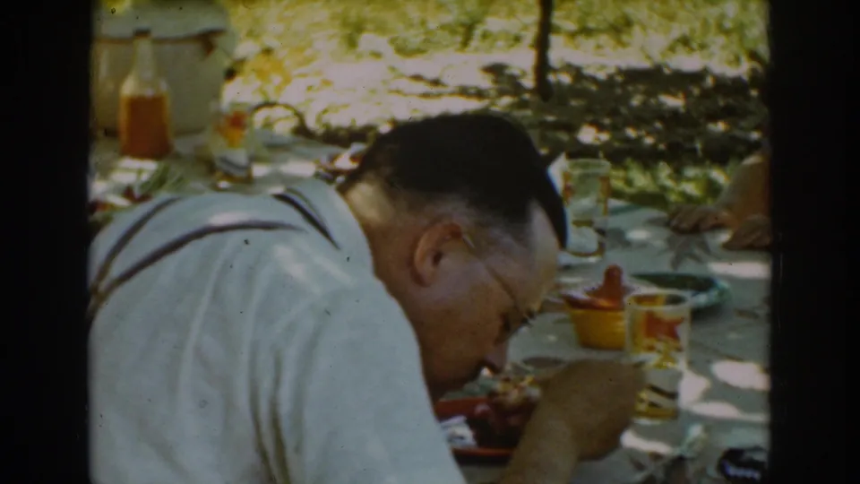 The final frame keeps focus on the man at the Auburn picnic table, with food, glasses, and another seated adult partly visible in the background.