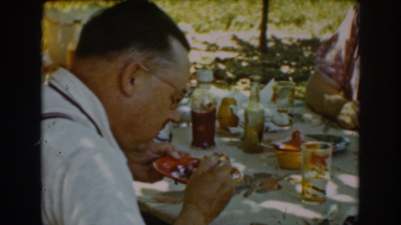 Man in glasses eating at a shaded outdoor table with plates and bottles.