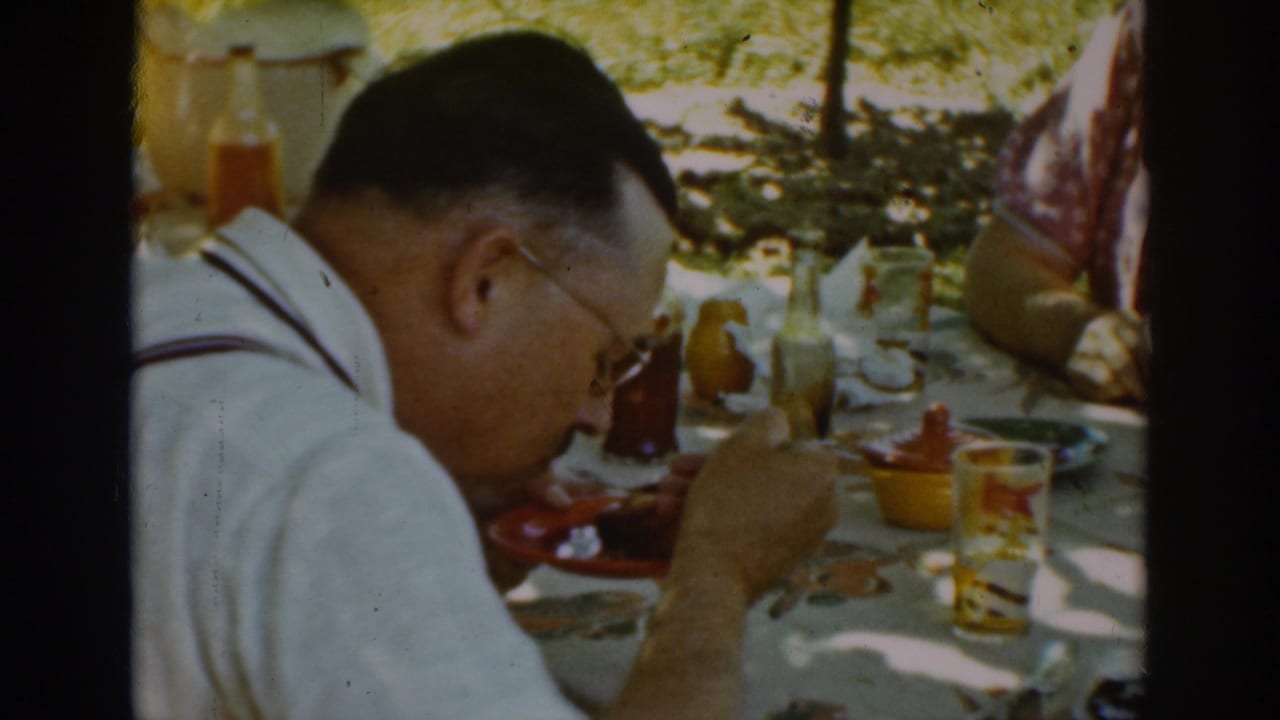 A 1939 Auburn home-movie frame shows a man seated in profile at a shaded outdoor table with plates, glasses, and condiment bottles.