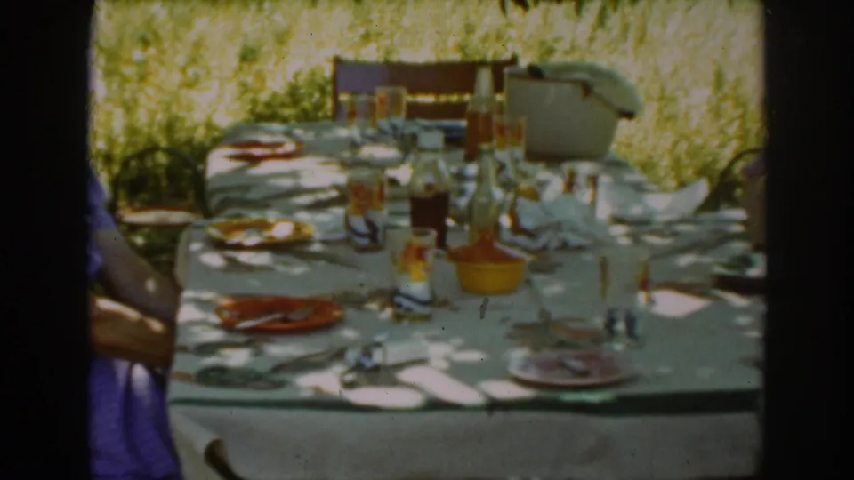 The camera lingers on the Auburn outdoor meal table, showing plates, bottles, glasses, a tablecloth, and a partially visible person at the edge.