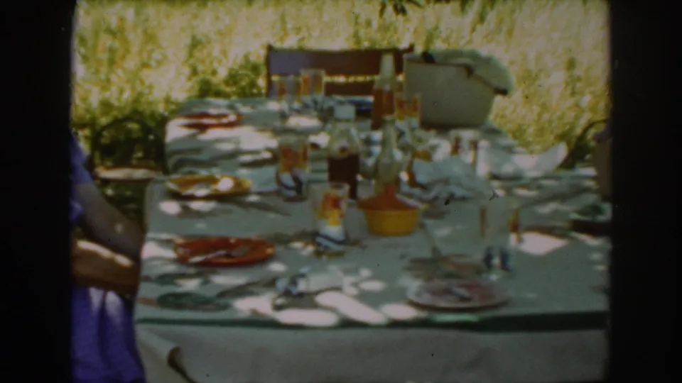A late-1930s Auburn outdoor dining table appears in soft 8mm color, with bottles, glassware, plates, and a partial person at left.