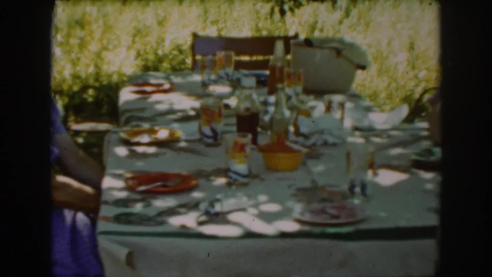 Sunlit Auburn picnic table in 1939 home-movie footage, with glassware, bottles, plates, and outdoor chairs under trees.