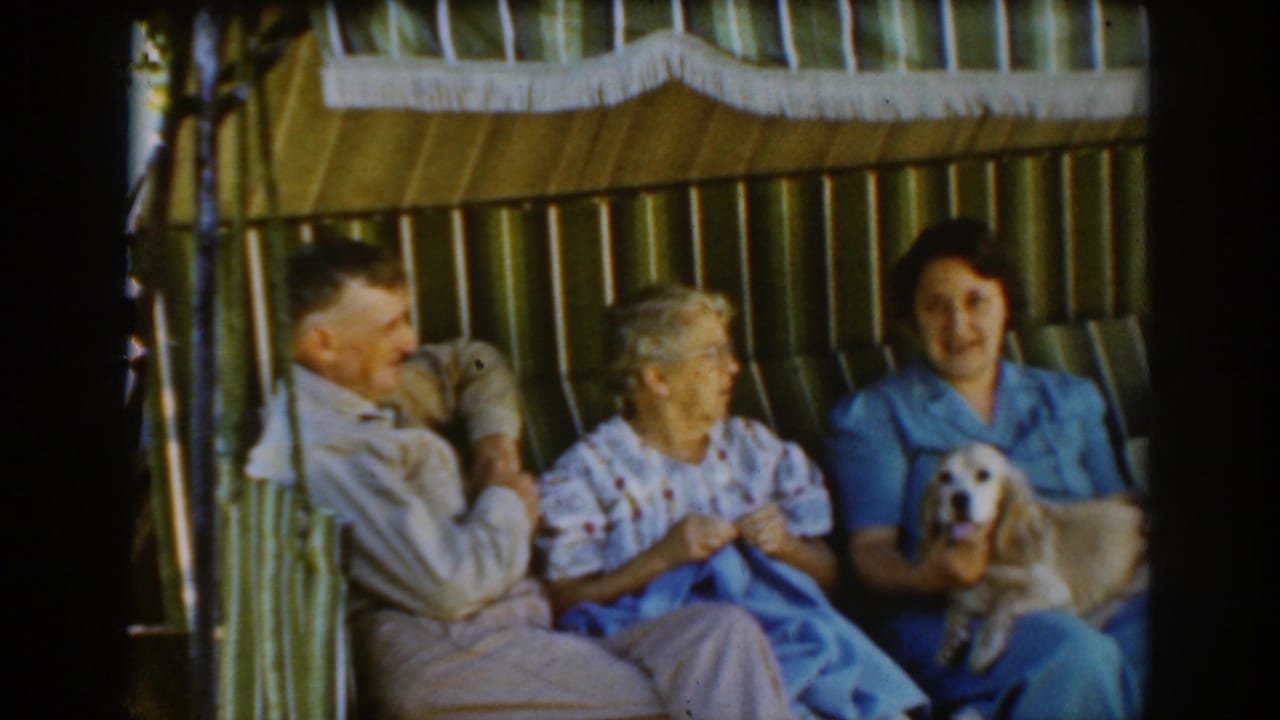 Three adults sit in a striped cabana while a younger woman holds a small light-colored dog.