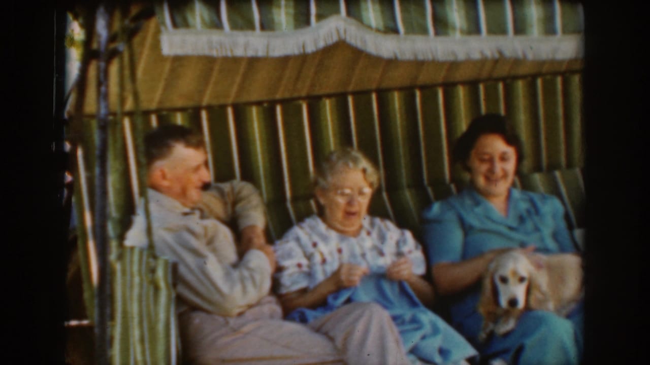 Three older adults sit on a striped outdoor swing, with one woman knitting and another holding a small dog.