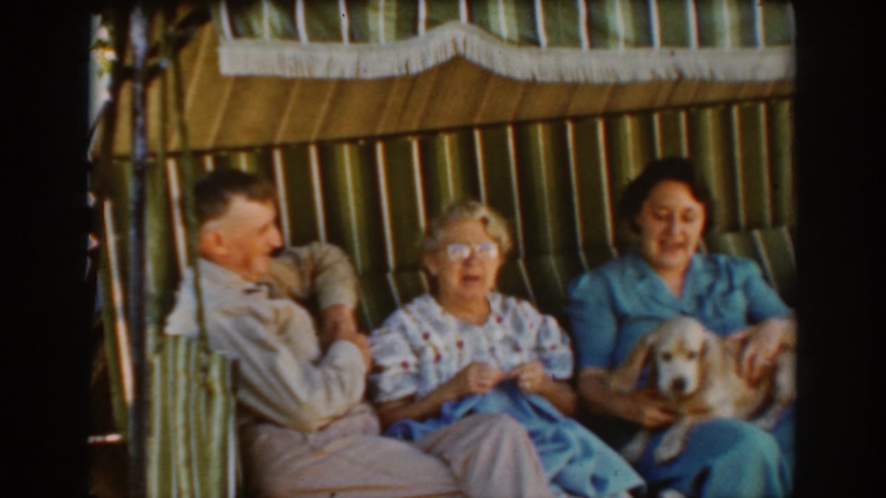 Auburn, California home-movie frame from 1939 showing three older adults seated on a striped swing, with the woman in blue holding a small dog.