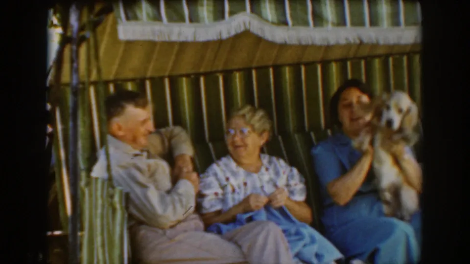 In the 1939 Auburn home movie, the woman in blue raises the small dog beside two older adults seated on a striped outdoor swing.