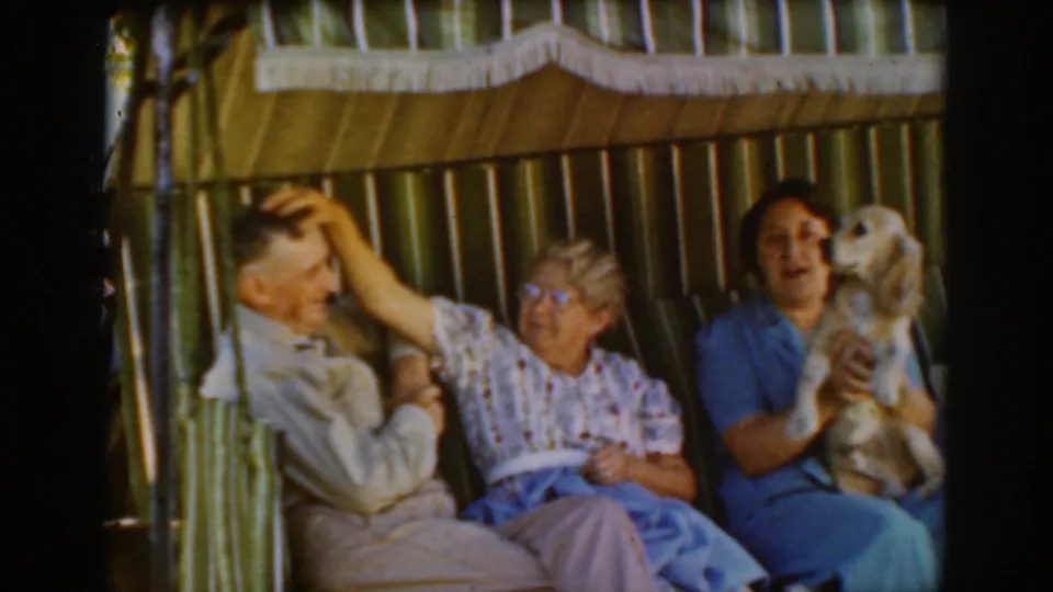 A 1939 Auburn home-movie frame shows an older woman reaching toward the seated man's head as a woman in blue holds a small dog beside them.