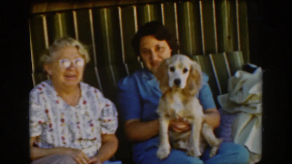 The older woman smiles while the woman in blue holds a light-colored spaniel facing the camera on the striped porch swing in 1939 Auburn footage.