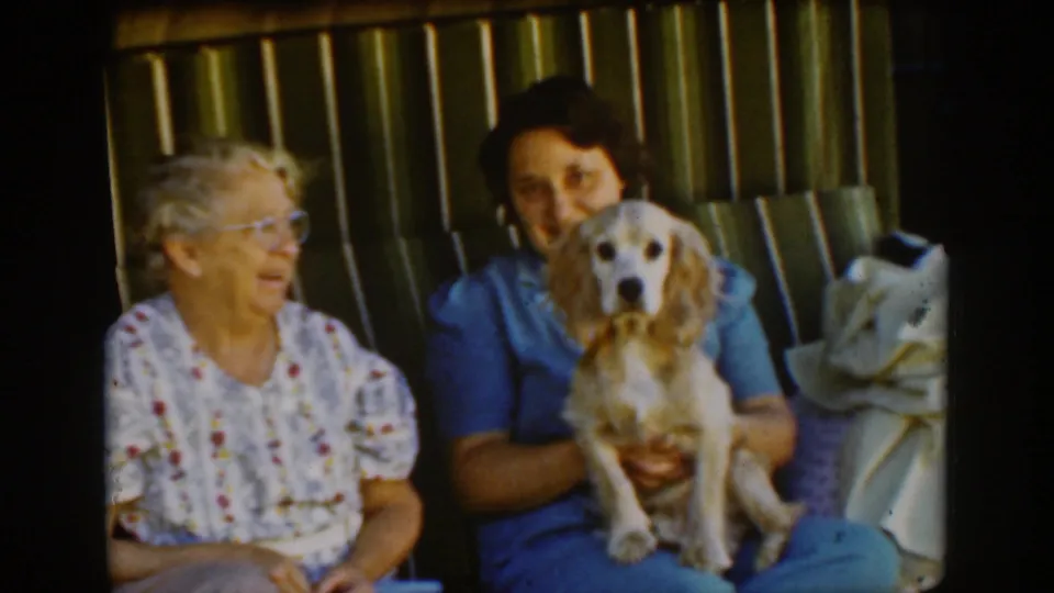 Two women sit on the striped porch swing in Auburn, California, with a light-colored spaniel held upright between them in the 1939 home movie.