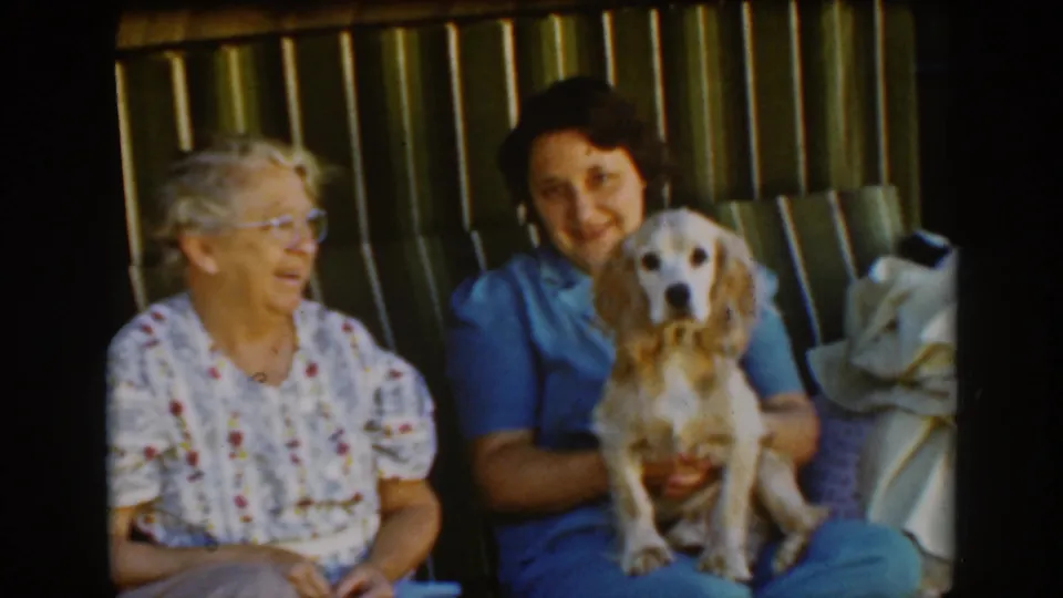 A 1939 Auburn home-movie frame shows the older woman turned toward the younger woman, who holds a small cocker spaniel on the bench.