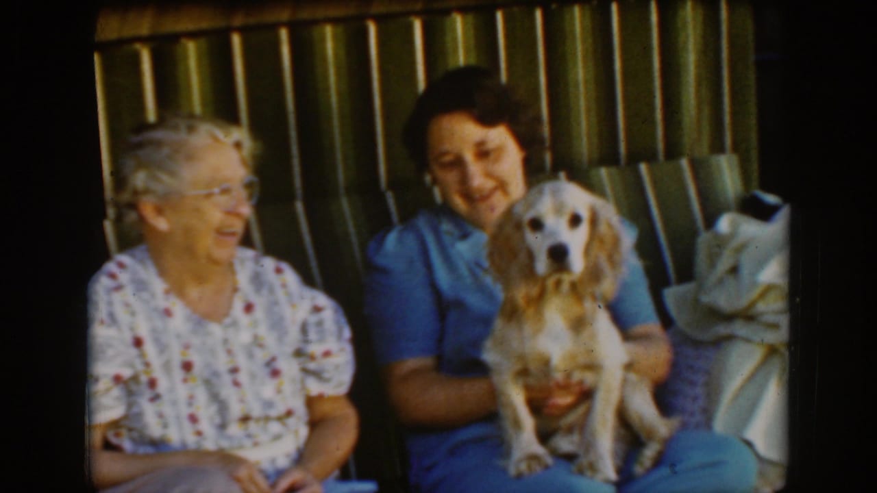 Two women sit on a porch as a dog faces the camera from the younger woman's lap.