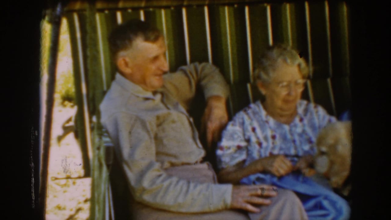 A man and older woman sit together on the Auburn porch as she handles knitting and the dog moves near her lap.