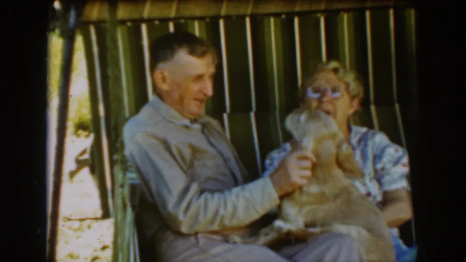 The elderly man lifts the tan cocker spaniel upright on the porch swing as the woman watches beside him in the Auburn home movie.