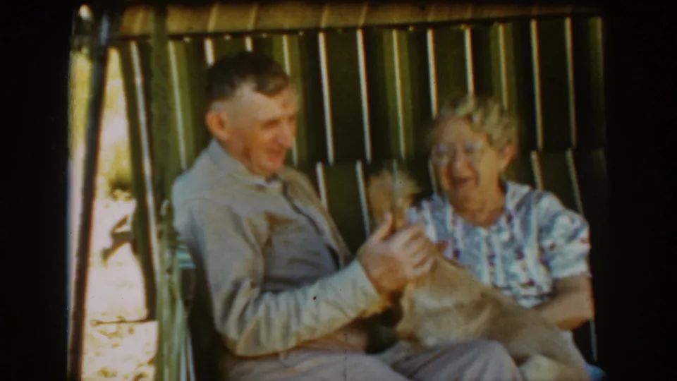The elderly couple appears amused as the tan dog moves on the man's lap in the 1939 Auburn porch-swing footage.