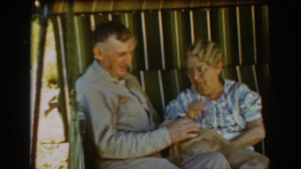 The woman gestures toward the dog as the elderly couple sits together on the porch swing in Auburn, California home-movie footage.