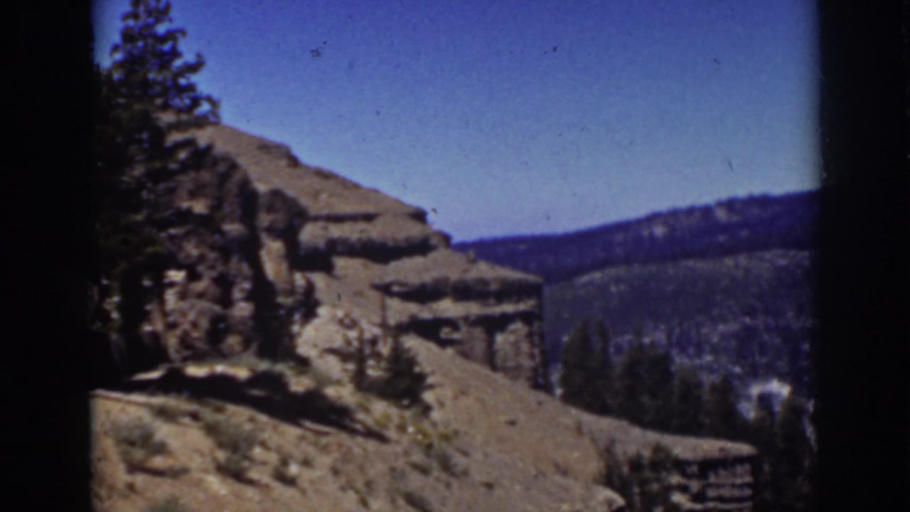 Rocky hillside with evergreen trees, distant forested ridge, and blue sky.