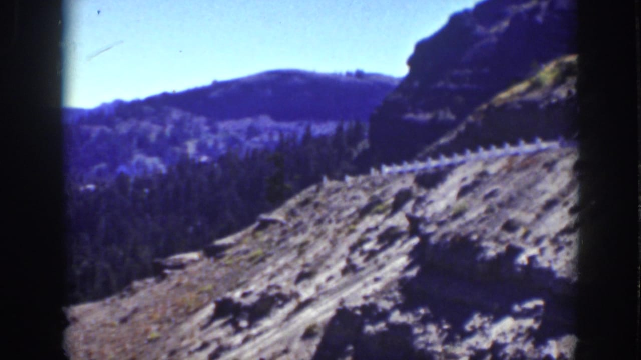 Rocky mountain slope with a dirt road and forested ridges beyond.