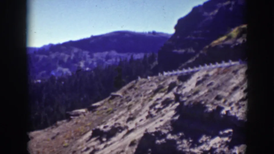 1939 Lake Tahoe frame with a rocky hillside in front, dark evergreen forest below, and a rounded distant ridge.
