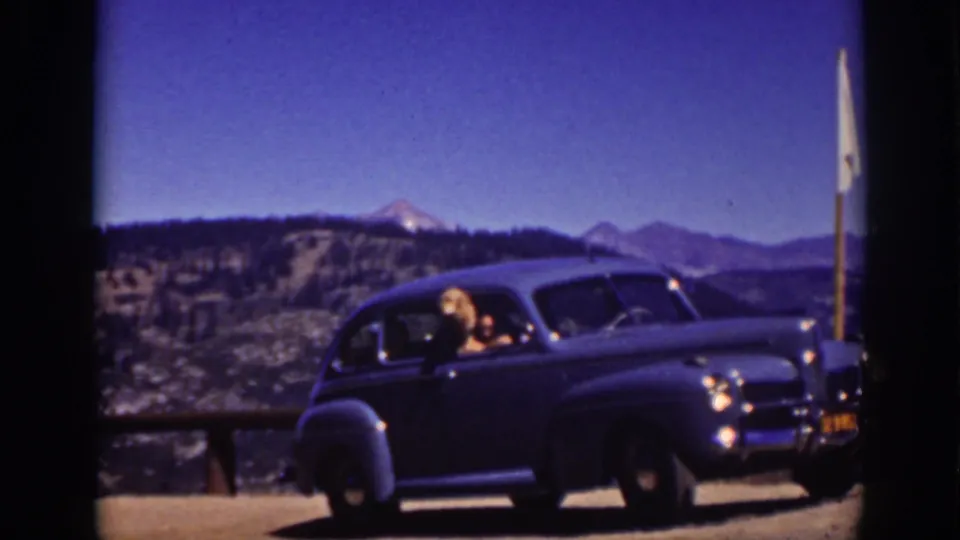 1939 Lake Tahoe home-movie frame with a woman at the passenger window of a dark classic sedan beside a roadside railing and mountain lake view.