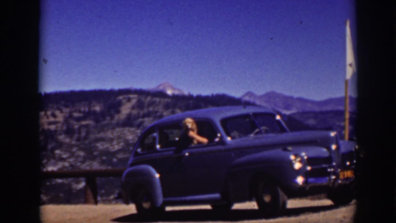1939 Lake Tahoe frame showing a dark rounded sedan stopped by a wooden rail as a woman looks out toward the camera with mountains behind.