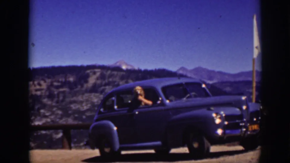 1939 Lake Tahoe frame showing a dark rounded sedan stopped by a wooden rail as a woman looks out toward the camera with mountains behind.