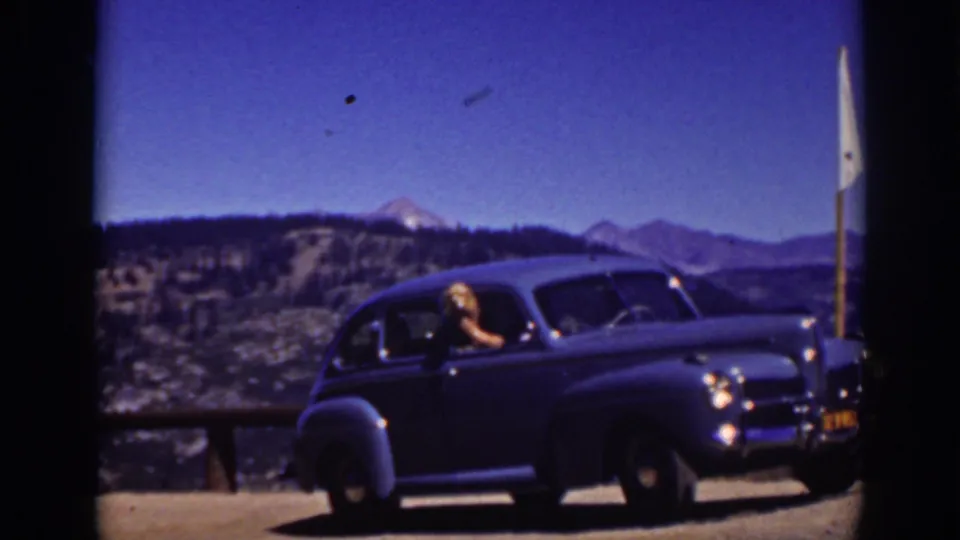 1939 Lake Tahoe frame of a woman in a dark classic sedan beside a wooden overlook rail, with lake water and mountain ridges in the distance.