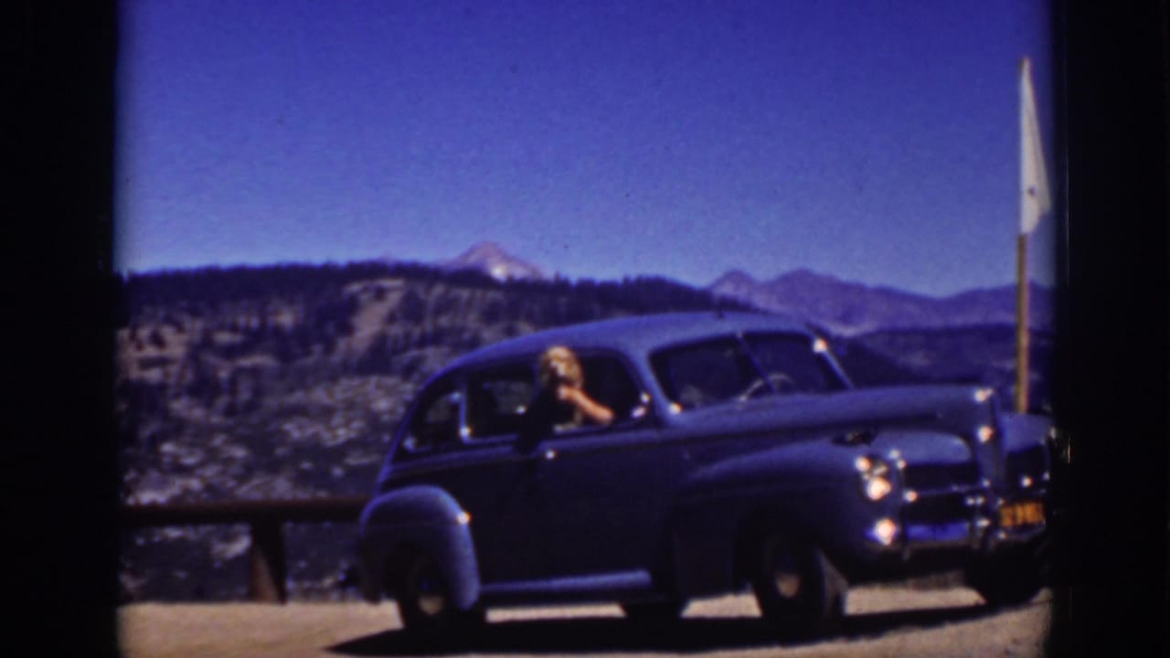 Blue vintage sedan parked at a mountain overlook with a blonde woman leaning from the driver's window.