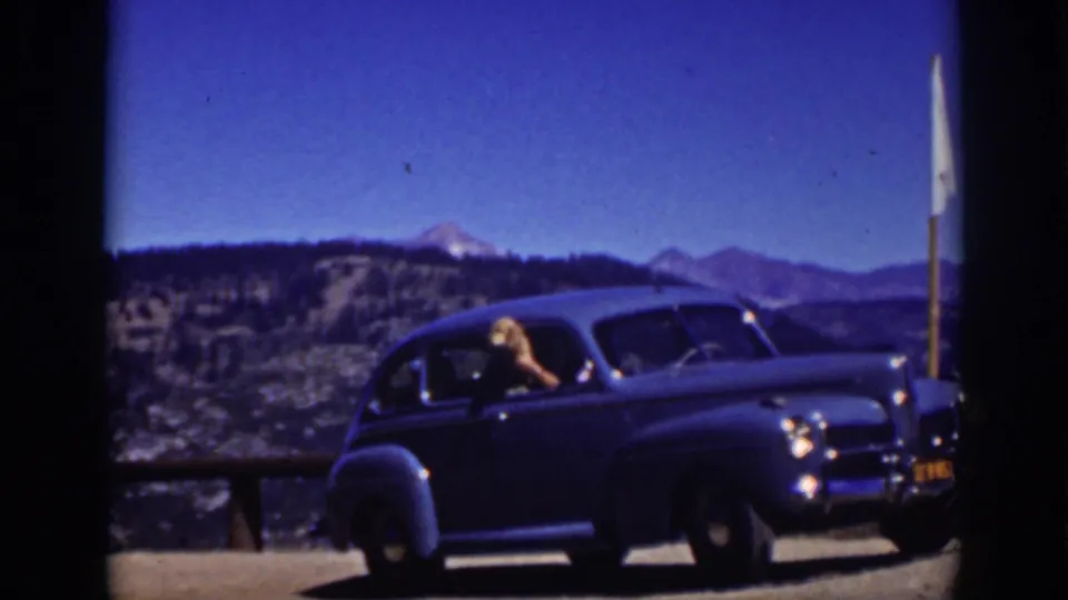 A 1939 Lake Tahoe travel frame showing the blue sedan from the side, the driver leaning out, and distant mountain peaks under bright sun.
