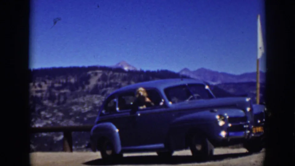 Final 1939 Lake Tahoe frame with the blue sedan still at the overlook, a woman visible in the driver's window, and hills beyond the water.