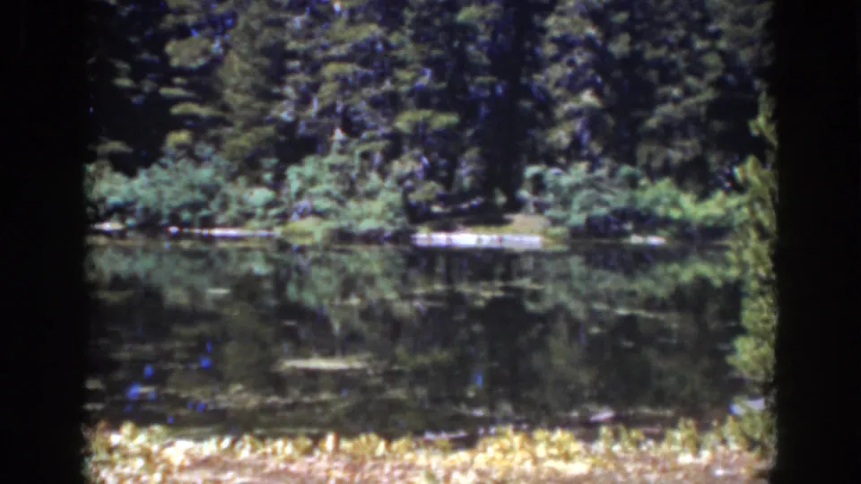 Lake Tahoe home-movie frame showing evergreen shoreline, still water reflections, and a small light dock or structure near the far bank.