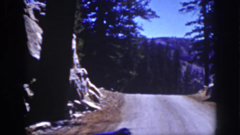 A Lake Tahoe road-view frame shows a pale gravel road bordered by conifers, rock formations, and distant forested hills.