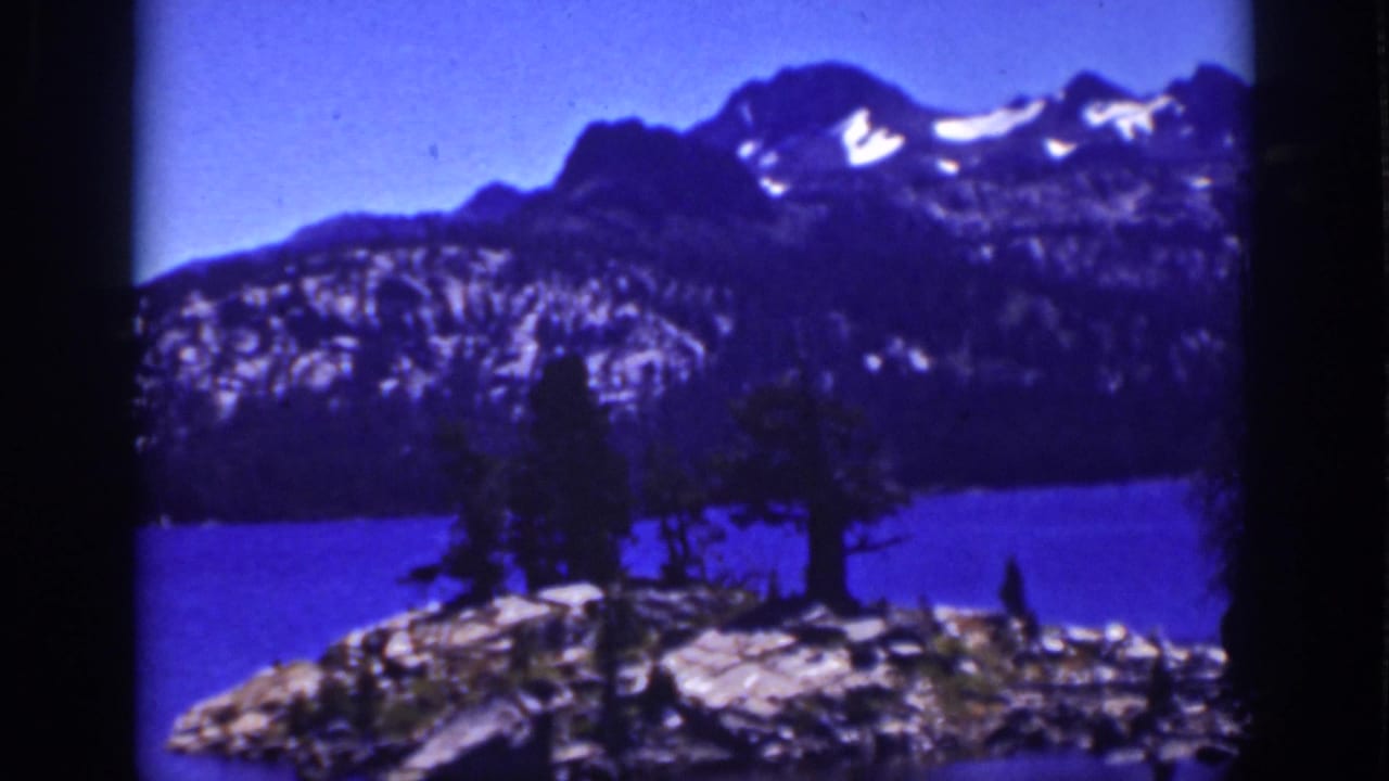 Blue alpine lake with rocky foreground, trees, and dark mountain peaks with snow patches.