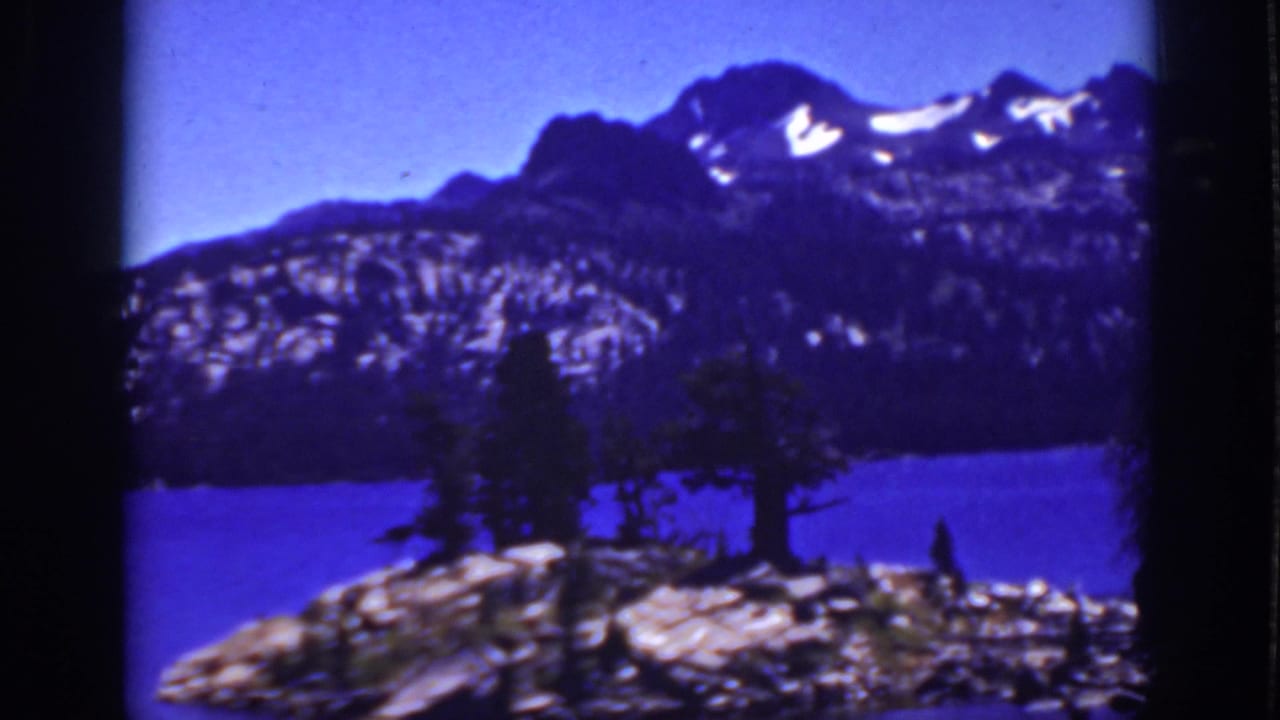 1939 Lake Tahoe-area frame with a small rocky, tree-covered shoreline or island set against a blue lake and mountain ridge.