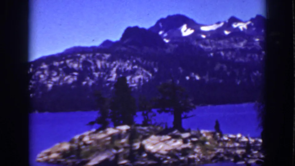 1939 Lake Tahoe-area frame with a small rocky, tree-covered shoreline or island set against a blue lake and mountain ridge.