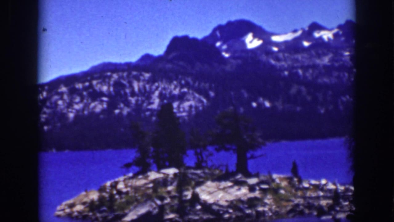 Lake Tahoe 8mm scenic frame showing trees on a rocky foreground, deep blue lake water, and snow-patched mountain peaks.