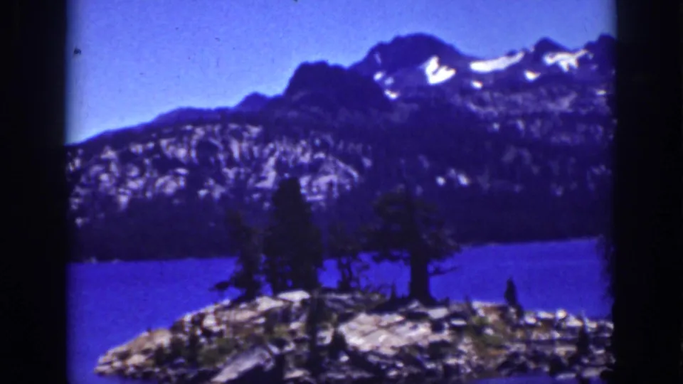 Lake Tahoe 8mm scenic frame showing trees on a rocky foreground, deep blue lake water, and snow-patched mountain peaks.