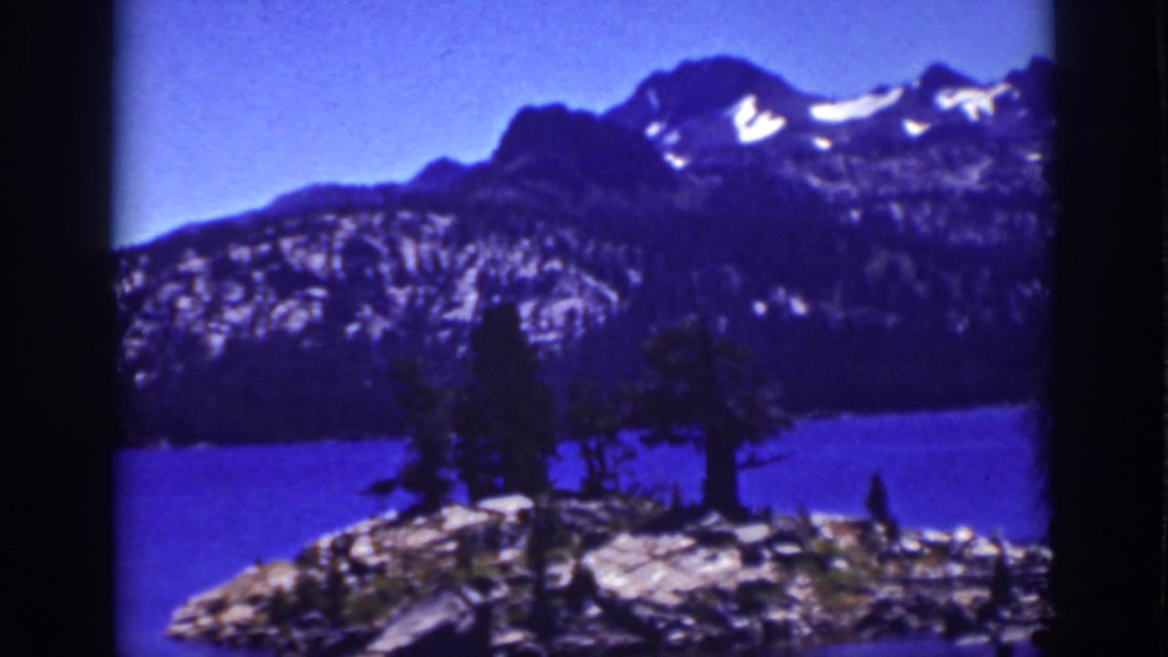 Mountain lake with rocky shoreline, pine trees, and snow-streaked peaks under a blue sky.
