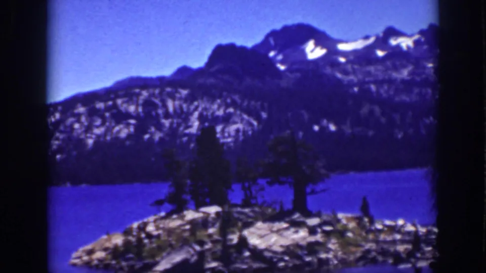 1939 8mm Lake Tahoe view with a blue lake, rocky foreground, pine trees, and snow-streaked Sierra Nevada peaks.
