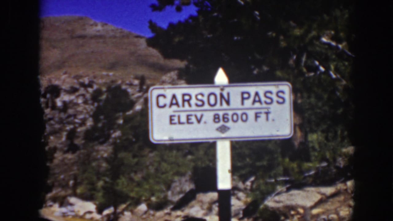 White road sign reading Carson Pass Elev. 8600 Ft. beside pines, rocks, and a rugged hillside in California travel footage.