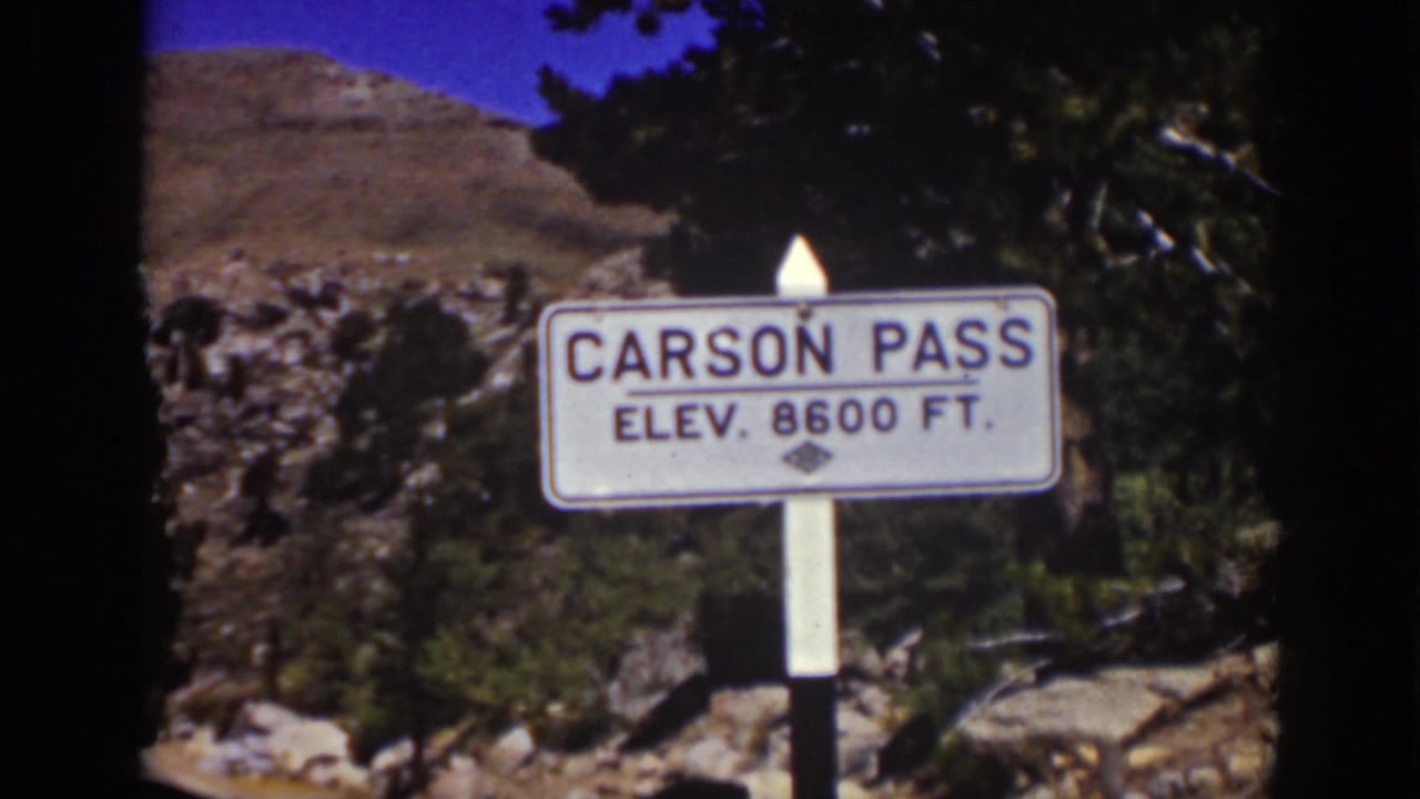1939 8mm frame of the Carson Pass 8600 ft sign with evergreen branches, a rocky hillside, and bright sky behind it.