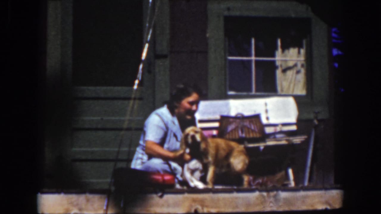 1939 home-movie frame from Lake Tahoe showing a smiling woman holding or washing a dog on a sunny wooden porch.
