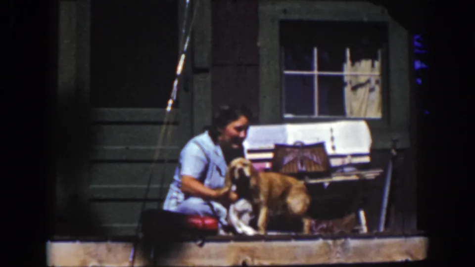 1939 home-movie frame from Lake Tahoe showing a smiling woman holding or washing a dog on a sunny wooden porch.