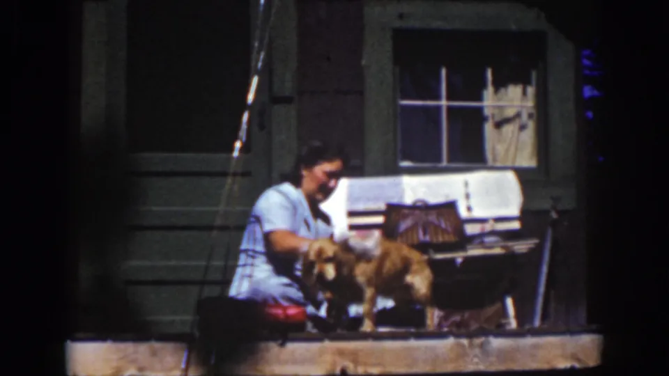 In 1939 Lake Tahoe home-movie footage, a seated woman in a light blue dress wipes a golden dog beside a porch window.
